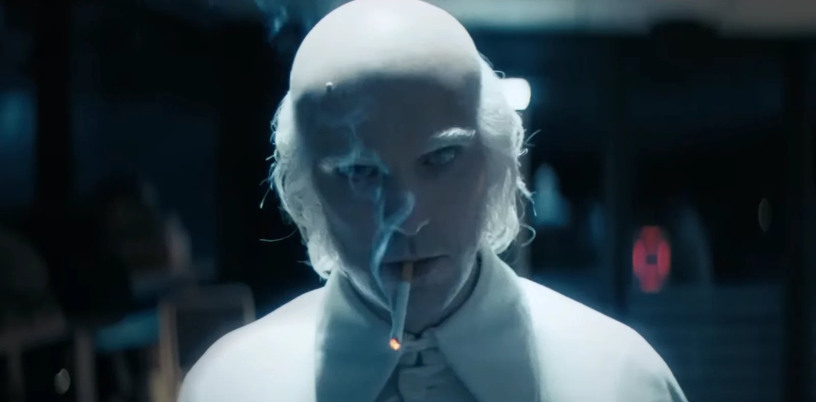A pale, bald man in a white suit smokes a cigarette in a dark laboratory-like setting.