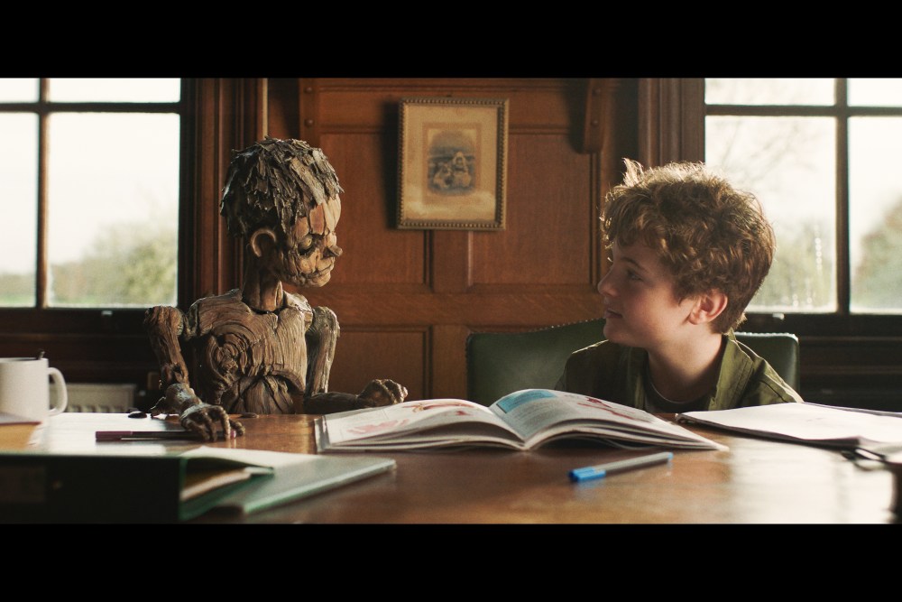 A young boy sits at a table beside a wooden puppet with an open book between them.