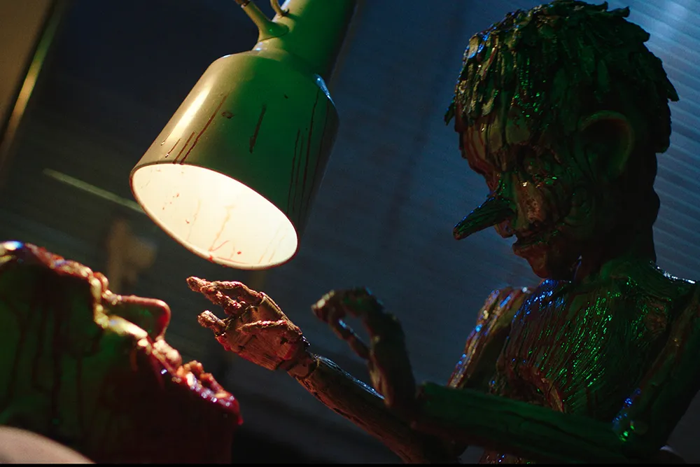 A blood-covered wooden Pinocchio reaches toward an injured person beneath a bright desk lamp.