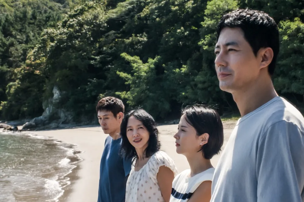 Four adults stand together on a beach with the sea and forest behind them