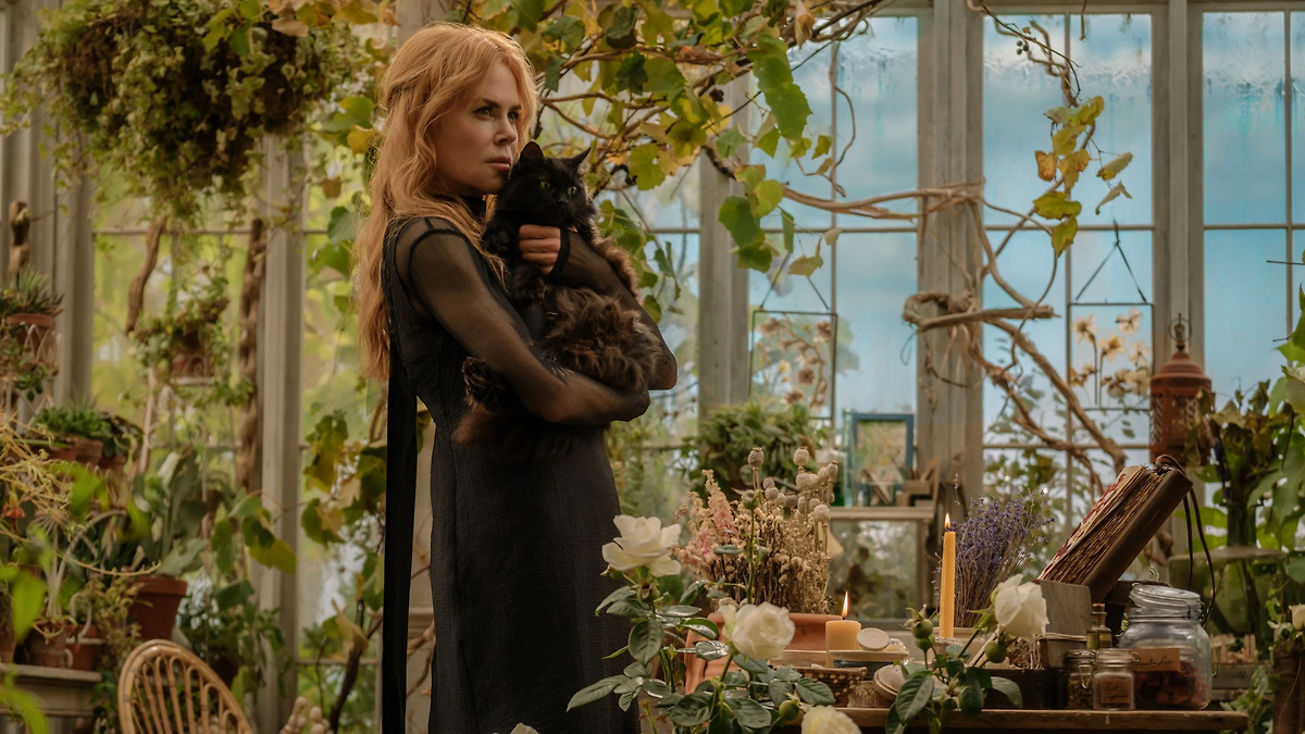 Nicole Kidman holds a black cat inside a sunlit greenhouse filled with plants, candles and books.