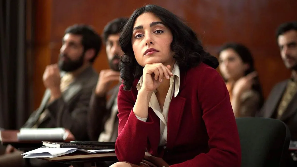 Golshifteh Farahani sits at a desk in a red cardigan while men listen behind her in a university setting.