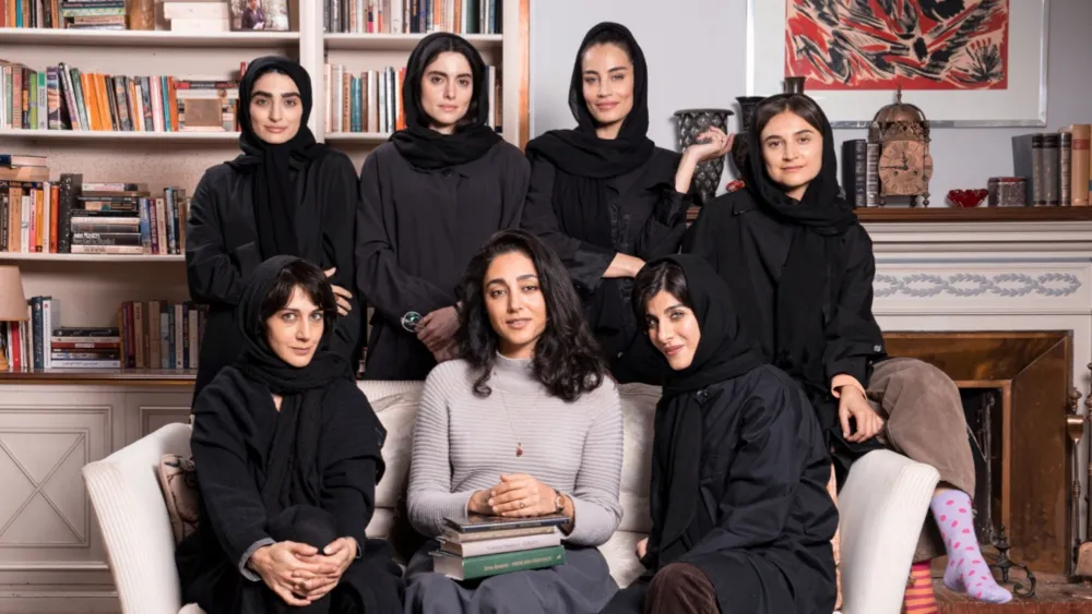 Seven women gather closely together in a book-lined room, several wearing black head coverings while others sit in front.