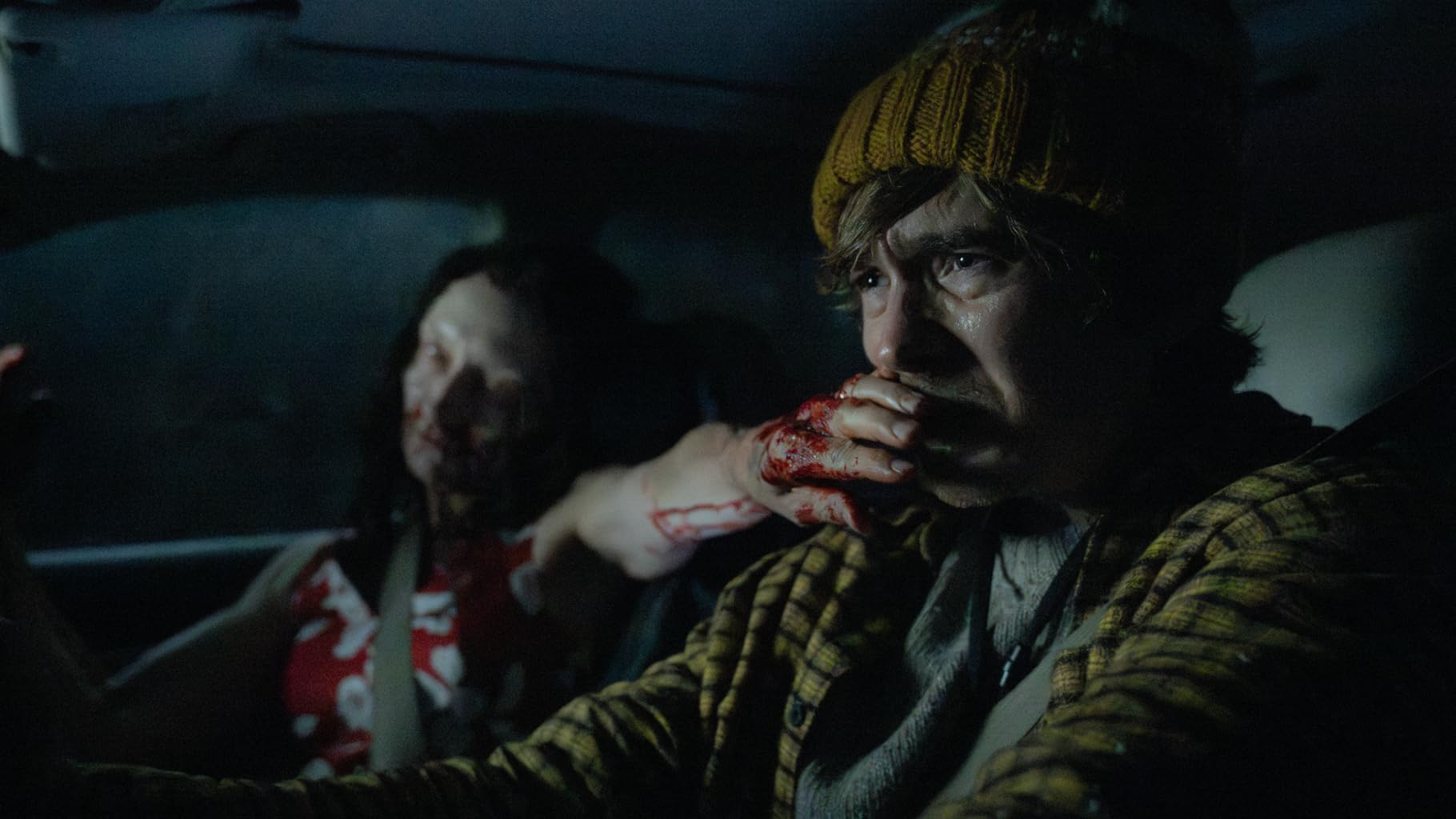 A bloodied man in a yellow knit cap drives through the dark while an infected passenger sits behind him.