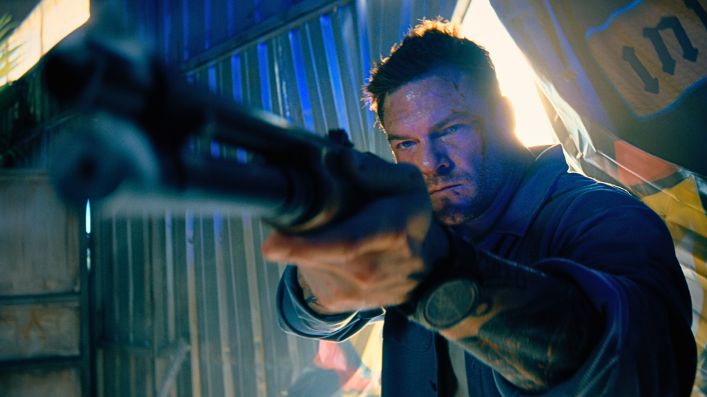 Alan Ritchson aims a shotgun inside a brightly lit industrial space.