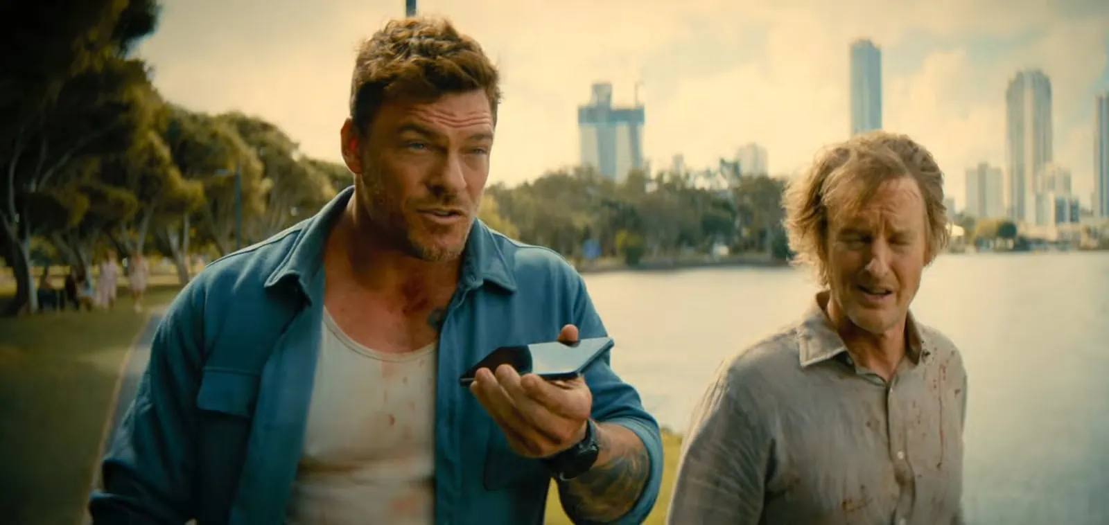 Alan Ritchson and Owen Wilson, both bloodied, hurry along a waterfront while speaking into a phone.