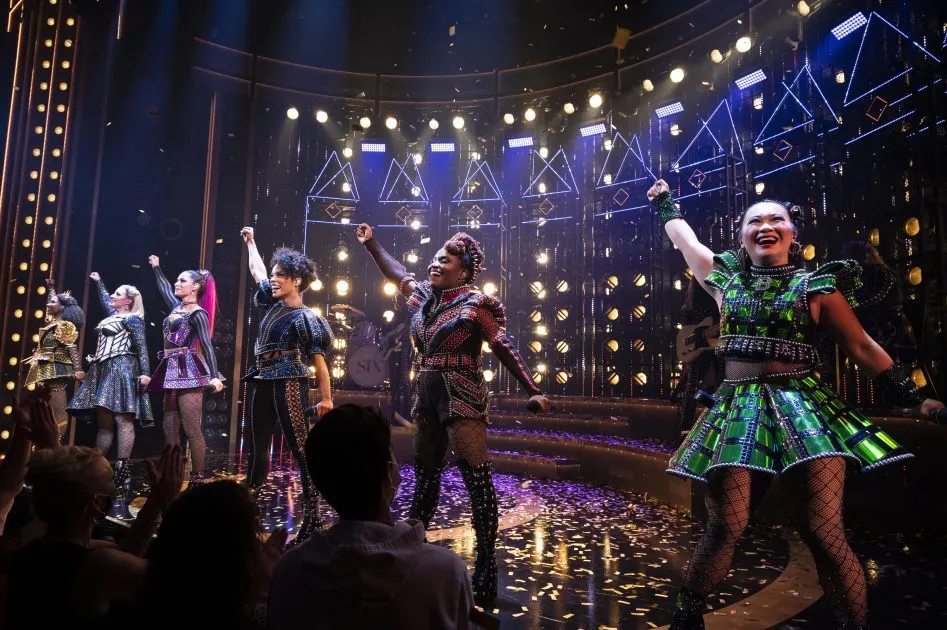 The six queens stand across the stage with their arms raised at the end of a performance of SIX.