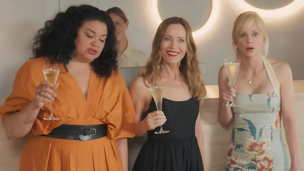 Michelle Buteau, Leslie Mann and Anna Faris hold drinks while talking inside a softly lit resort in Spa Weekend.
