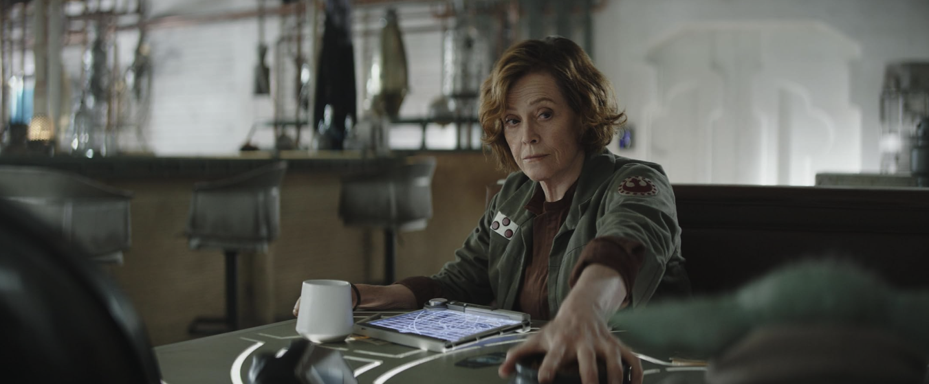 Sigourney Weaver sits at a table in a military operations room.