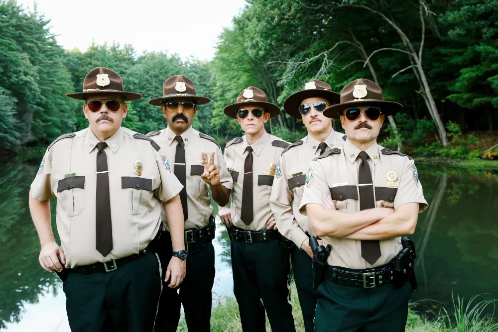 Five Vermont state troopers wearing sunglasses and campaign hats stand together beside a wooded pond.
