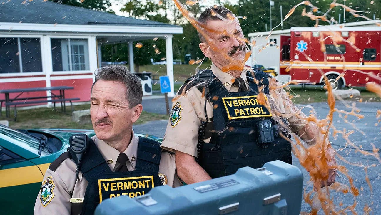 Two Vermont state troopers stand beside a patrol car as brown liquid sprays across one officer’s face and uniform.
