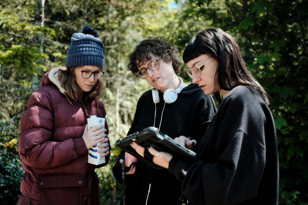 Three people stand outdoors around a production monitor surrounded by trees.