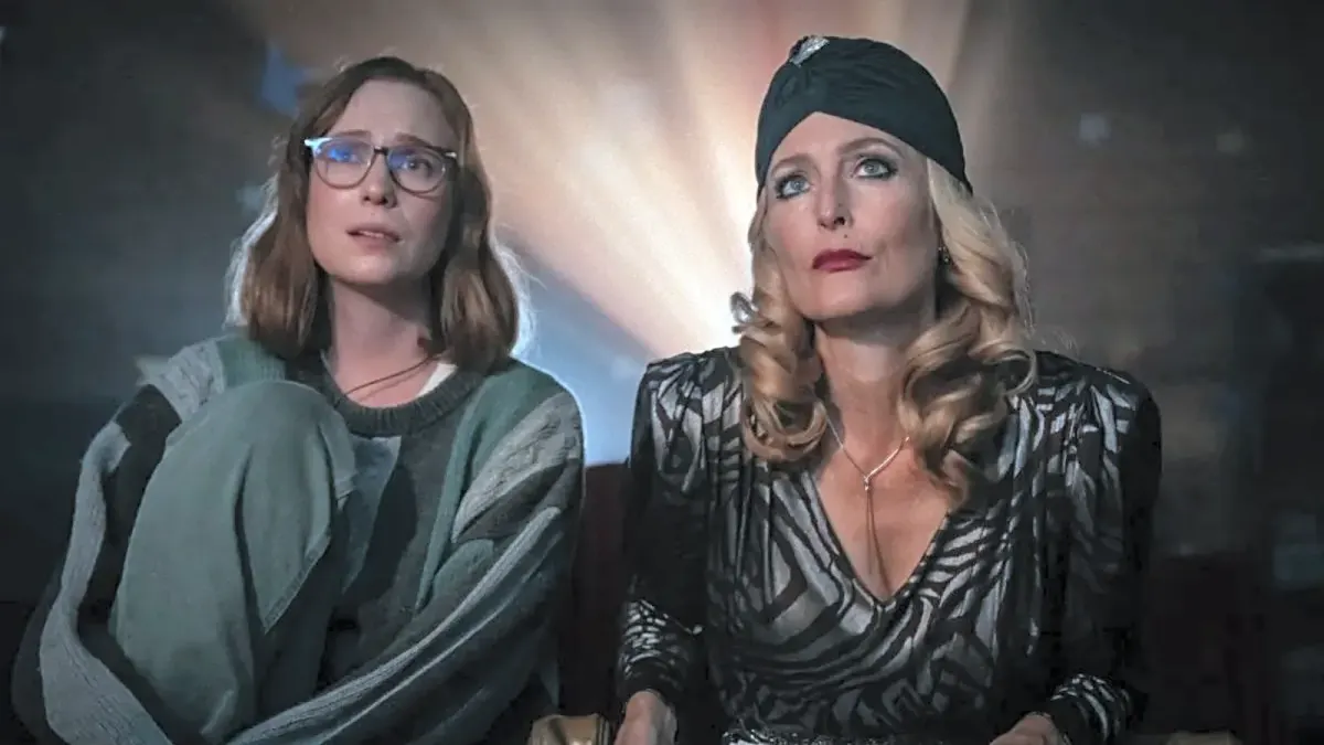 Two women sit beside each other in a dim room, one wearing glasses and the other dressed in a dark patterned outfit.