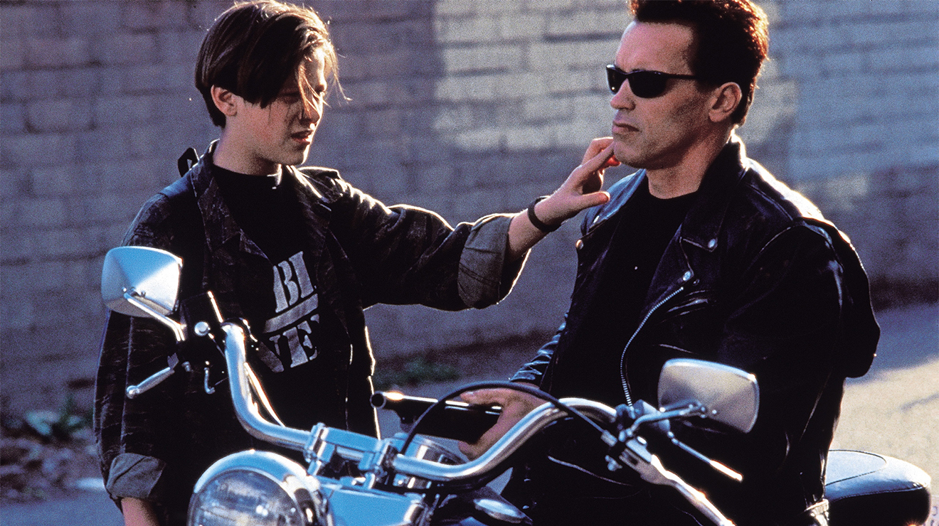 Edward Furlong as John Connor reaches toward Arnold Schwarzenegger’s T-800 beside a motorcycle.