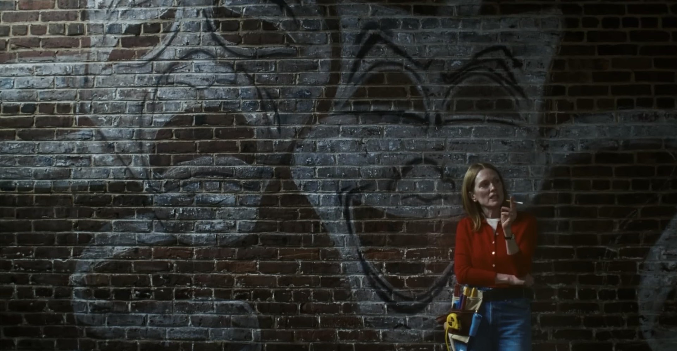 Julianne Moore stands in front of a graffiti-covered brick wall in costume with a work belt.