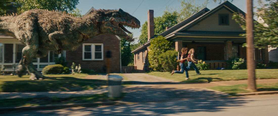 A dinosaur charges through a suburban neighborhood as two people run across a lawn in The End of Oak Street.