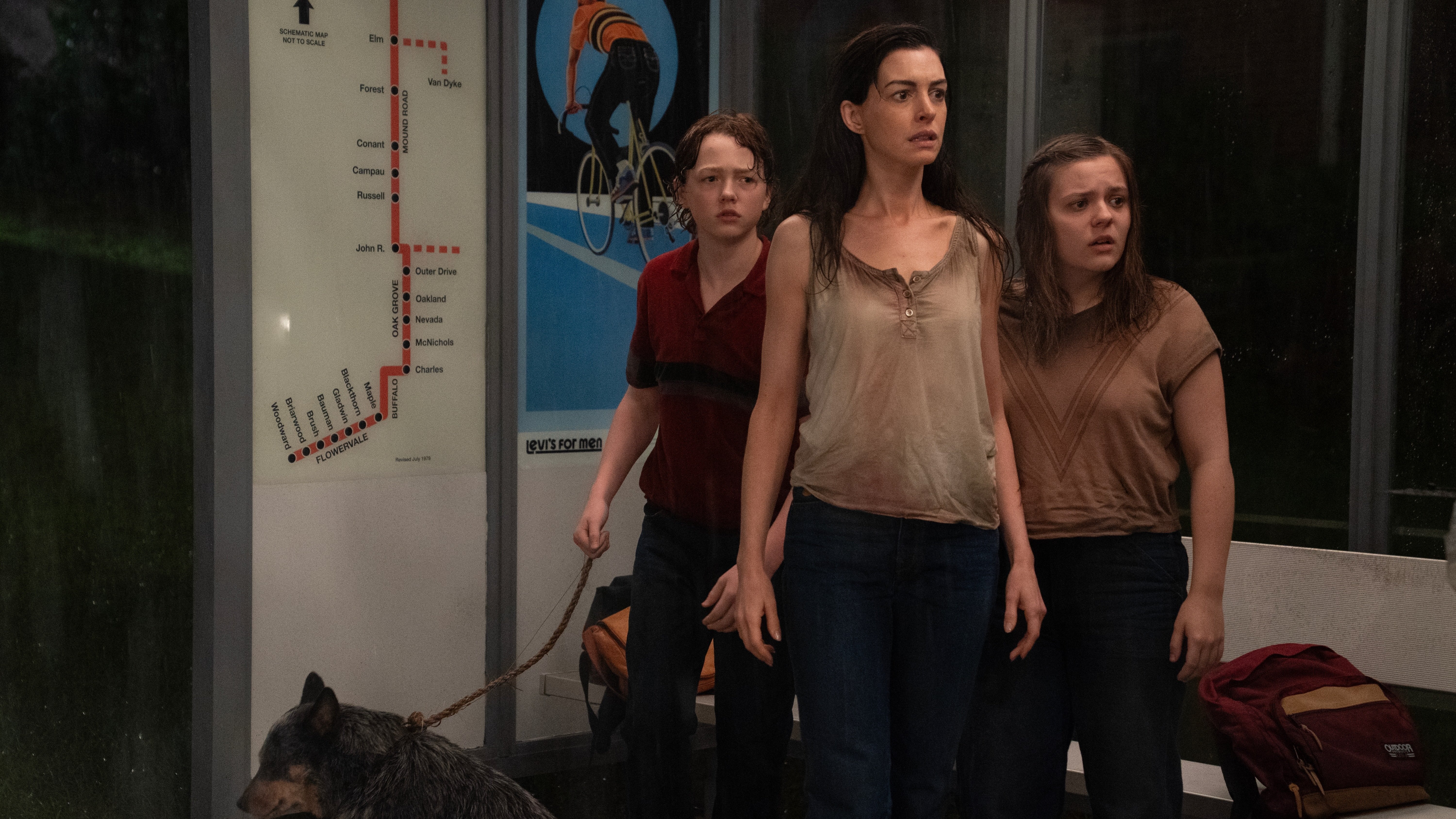 Anne Hathaway stands with two younger characters and a dog inside a bus shelter in The End of Oak Street.