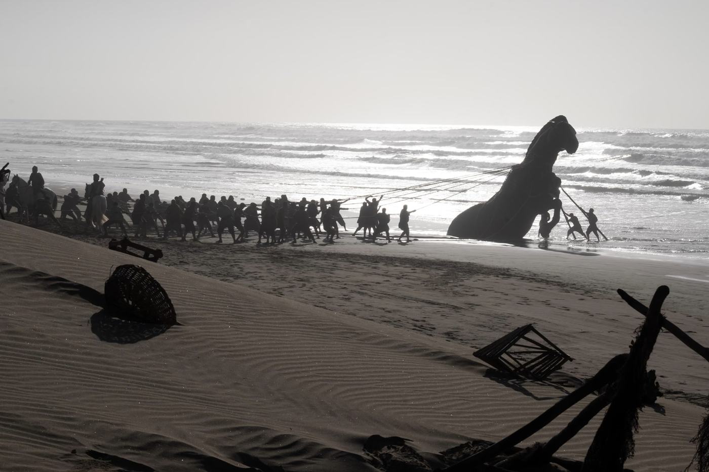 A massive wooden Trojan Horse lies partly submerged near the shoreline as soldiers haul it from the surf with ropes.