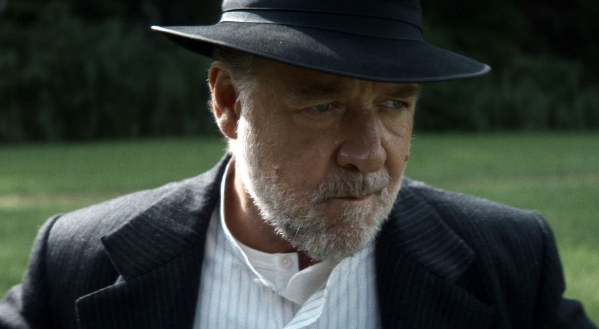 Russell Crowe, wearing a dark brimmed hat and pinstriped jacket, looks to the side in a sunlit field.