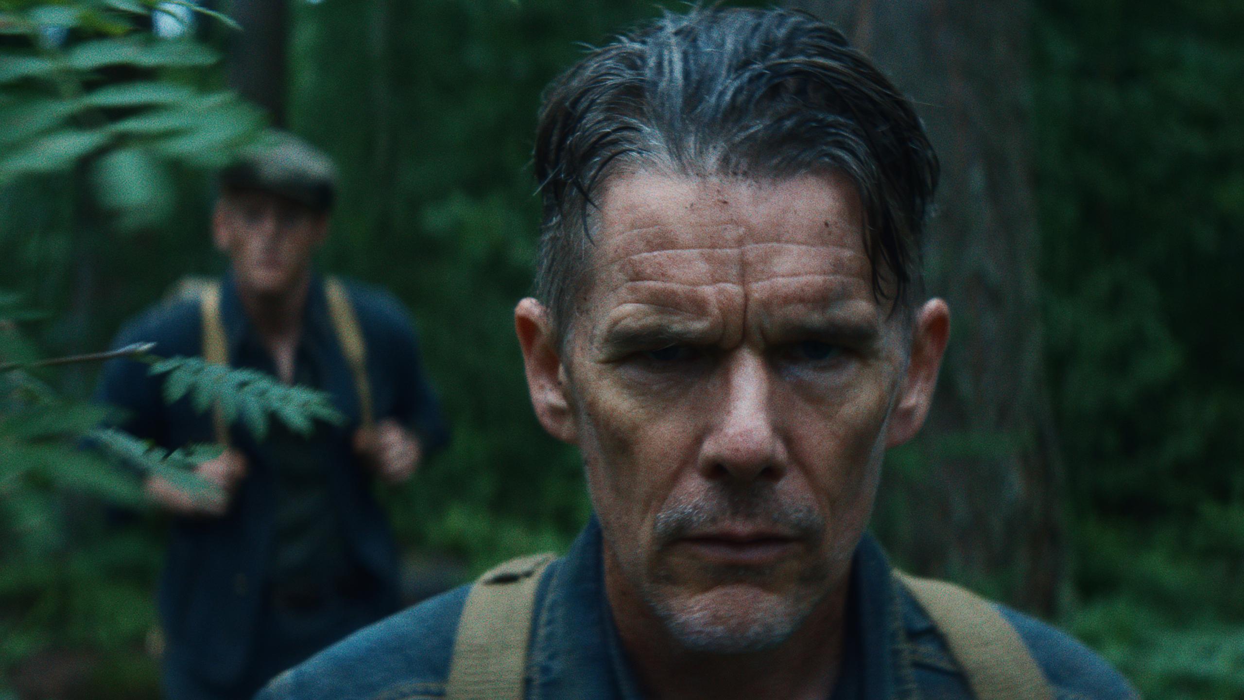 A wet and exhausted Ethan Hawke stands in a dense forest with another man blurred behind him.