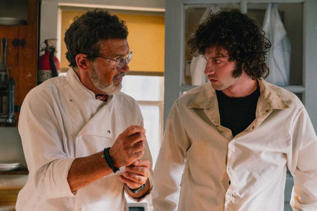 An older chef and a young kitchen worker in white uniforms stand talking beside each other in a restaurant kitchen.