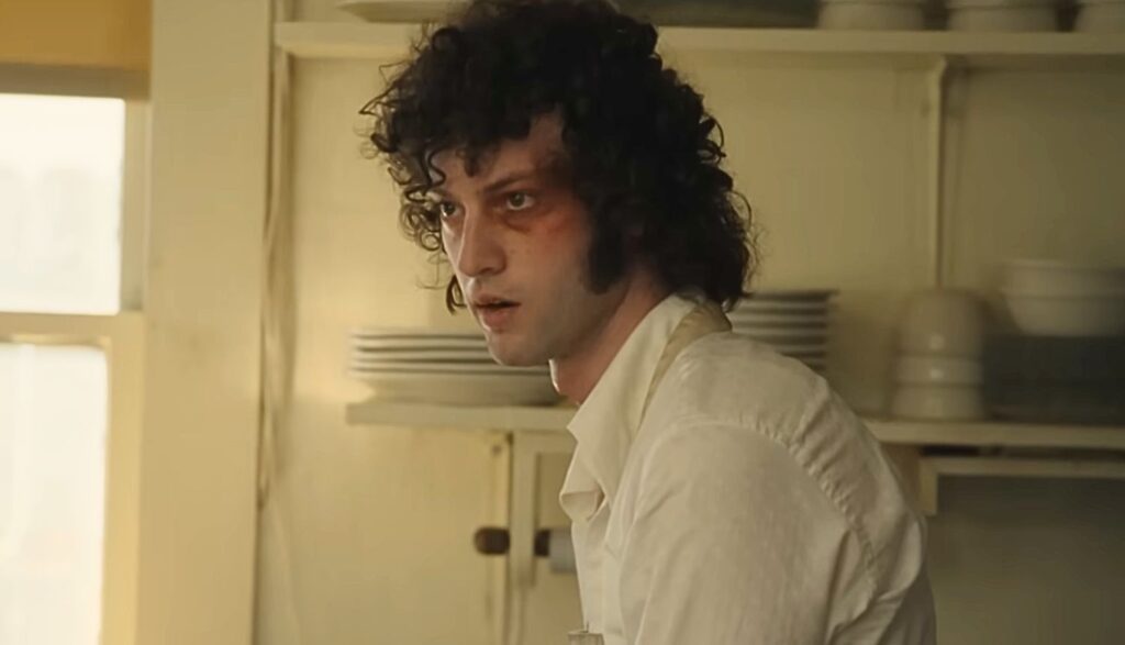 A young man with curly dark hair and a bruised eye stands alone in a restaurant kitchen wearing a white work shirt.