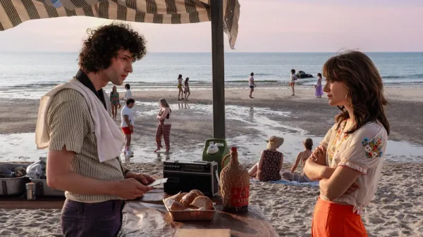 A young man working behind an outdoor food counter speaks with a young woman beside a crowded beach.