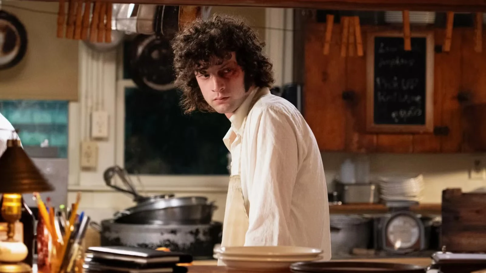A young man in a white work shirt looks across a restaurant kitchen with a bruise around one eye.