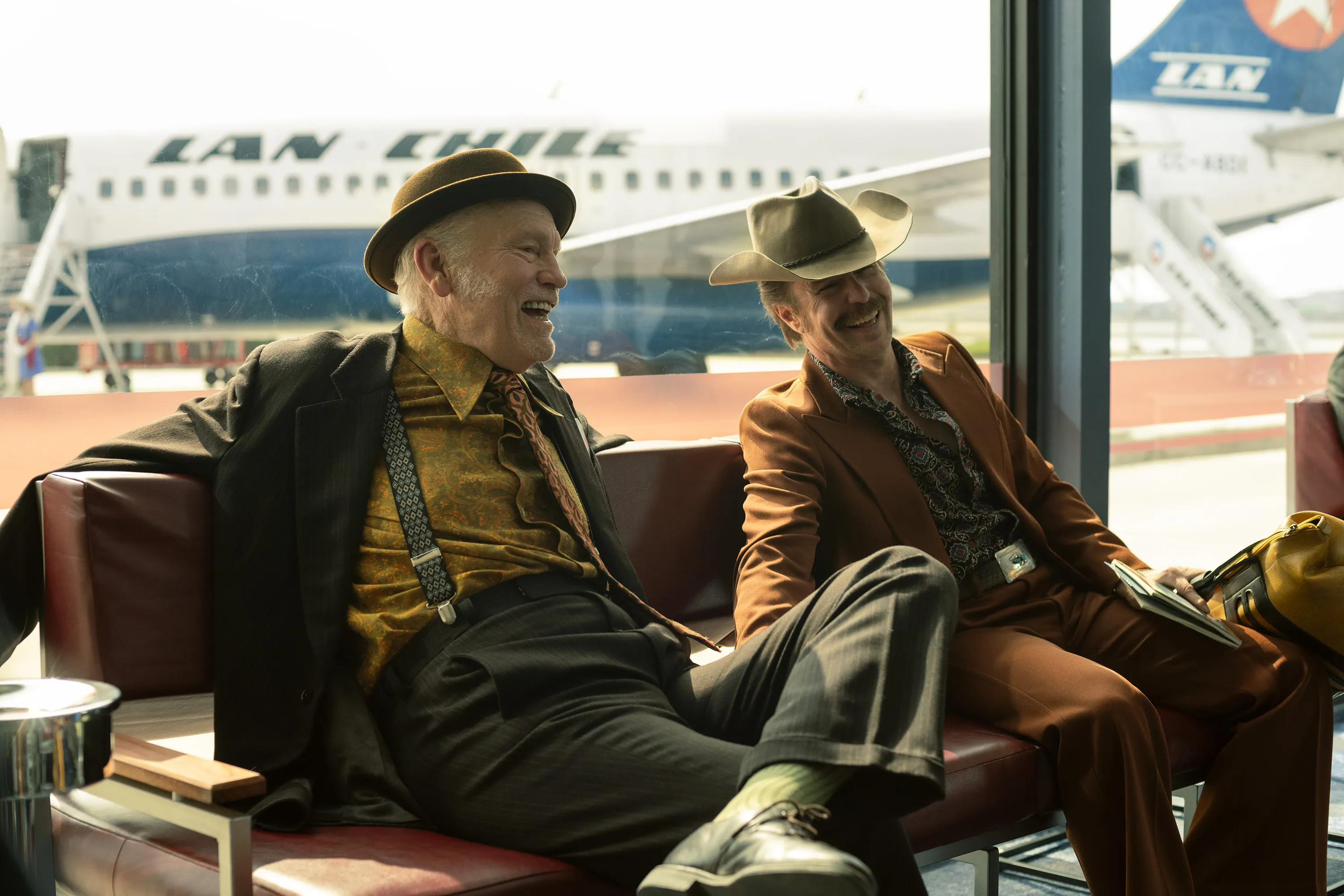 John Malkovich and Sam Rockwell sit together in an airport lounge, laughing beside a large passenger airplane.