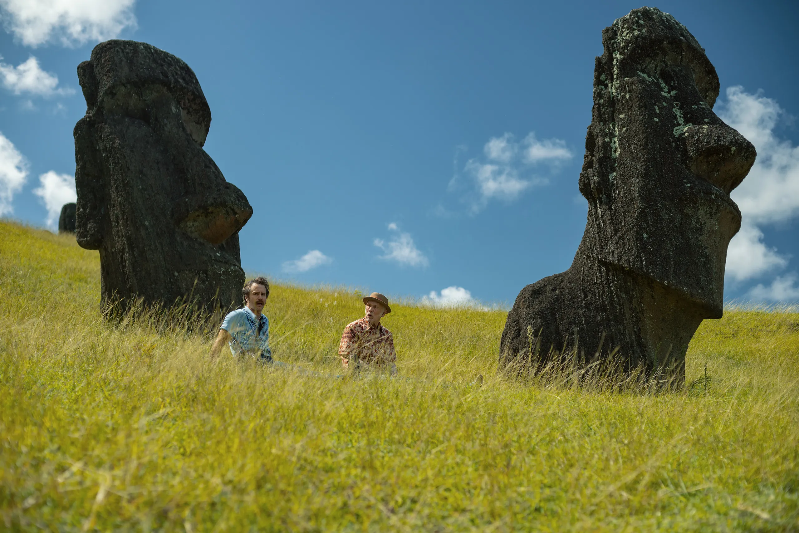 Sam Rockwell and John Malkovich walk through tall grass between the moai statues of Easter Island.