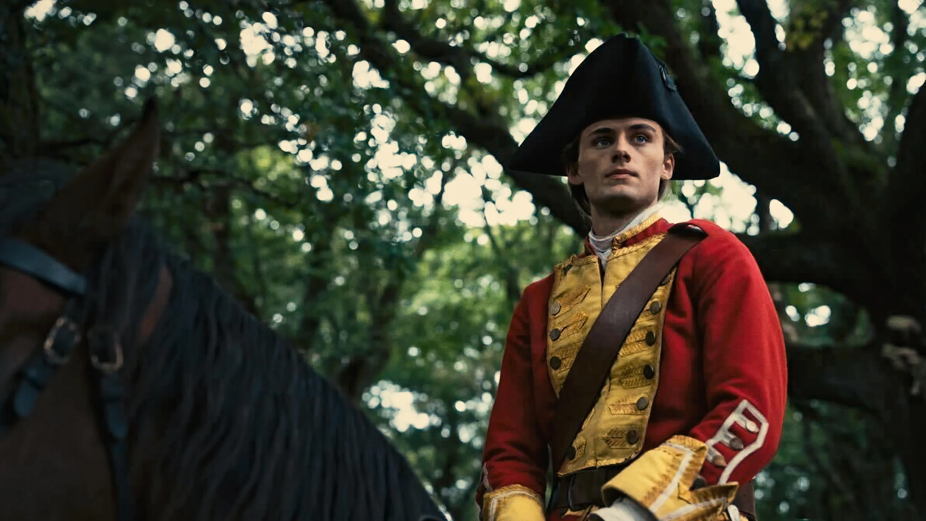 A young British officer in a red and gold eighteenth-century uniform sits on horseback beneath a canopy of trees.