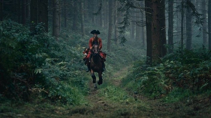 A young British officer in a red uniform rides a horse along a narrow path through a dense, misty forest.