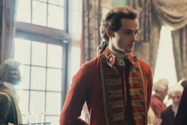 A young man in a red eighteenth-century military uniform stands in a bright formal room surrounded by colonial officials.